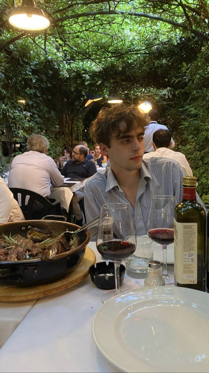 a young man sitting at a table with wine and food in front of him, surrounded by greenery