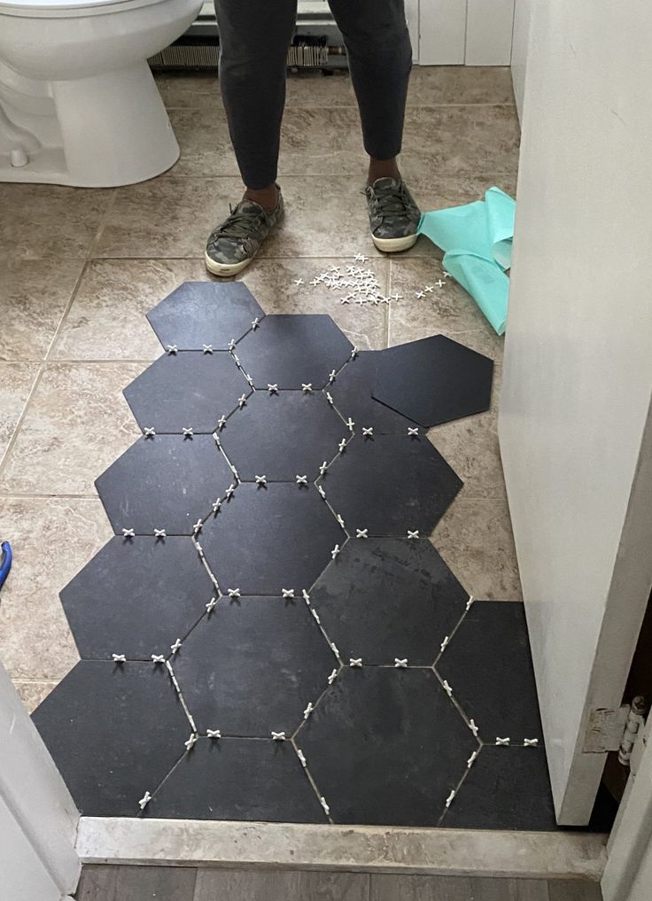 a person standing in front of a toilet on a bathroom floor with black hexagonal tiles