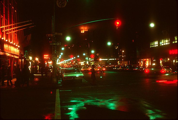 a city street at night with traffic lights and people walking on the sidewalk in the rain