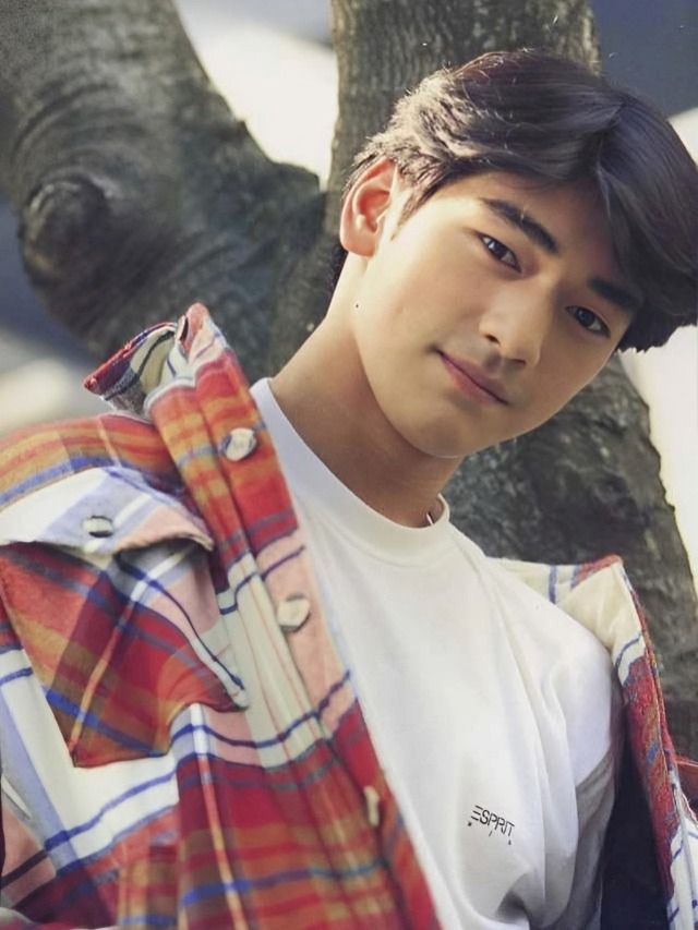 a young man standing in front of a tree wearing a plaid jacket and white t - shirt