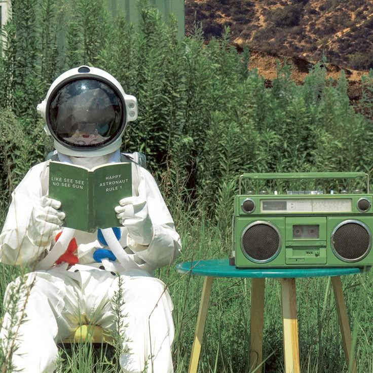 an astronaut reading a book while sitting on a chair in the grass next to a radio