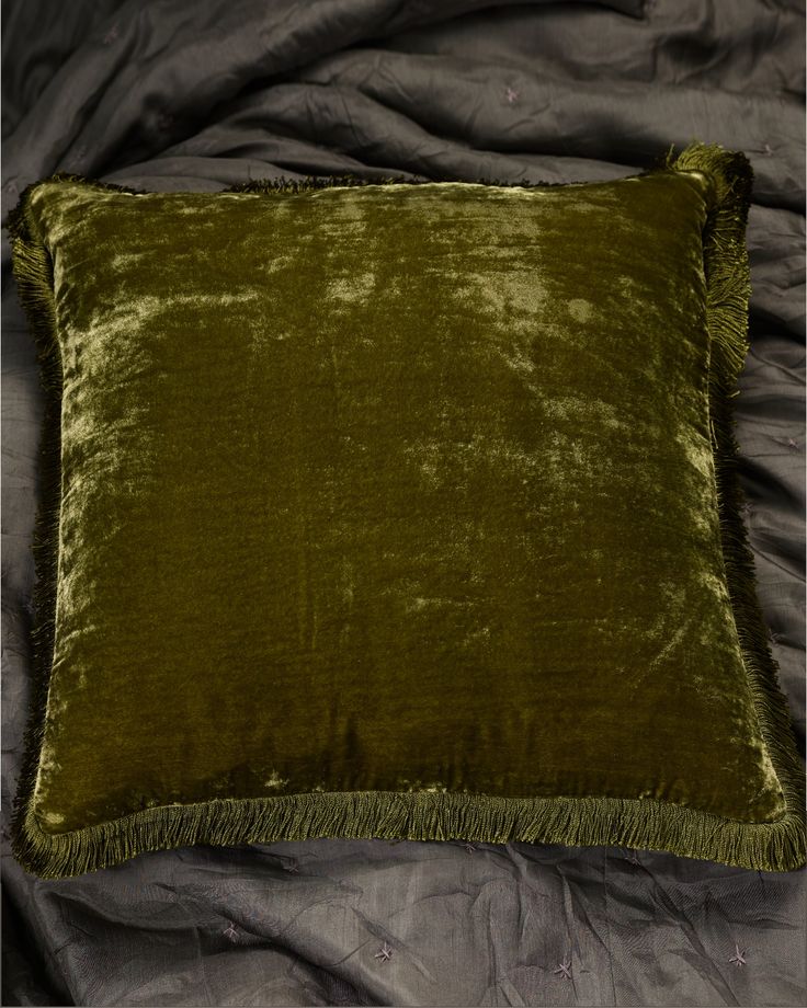 a green velvet pillow with fringe trim on a gray bed sheeted surface, top view