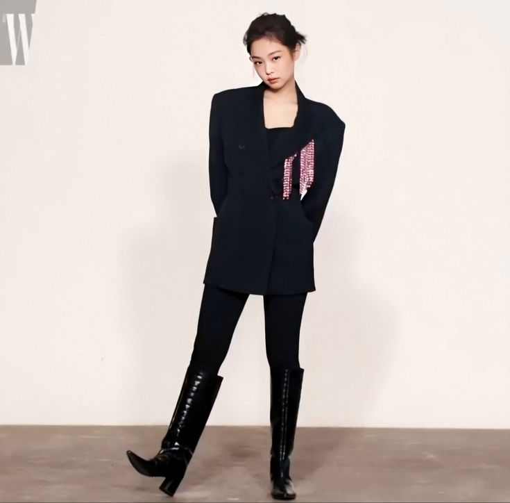 a woman standing in front of a white wall wearing black boots and a blazer