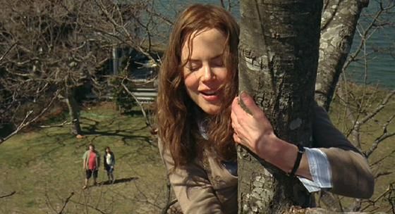 a woman standing next to a tree in front of a body of water with people walking by