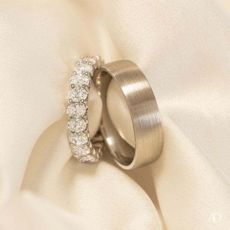 two wedding rings sitting on top of a white satin covered cloth with diamonds in the middle