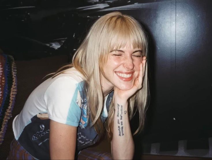 a woman with blonde hair smiling and leaning against a wall in front of a piano