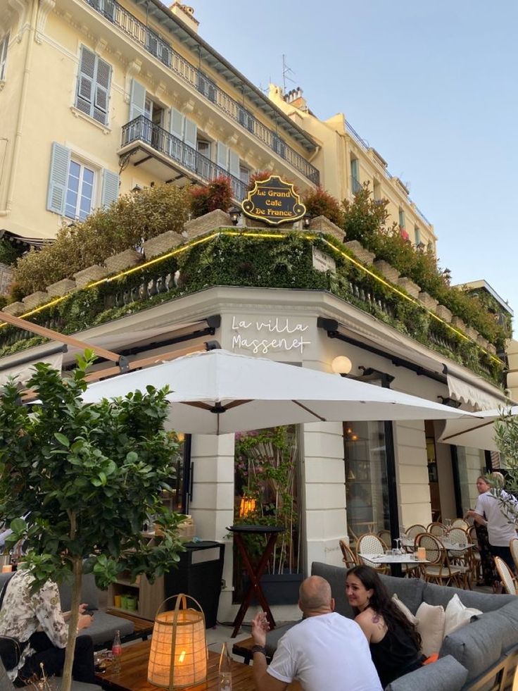 people sitting at tables in front of a building with plants growing on the top of it