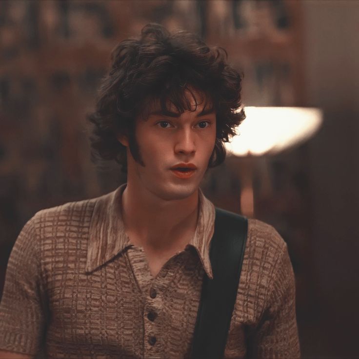 a young man with curly hair wearing a brown shirt and carrying a black bag looking at the camera