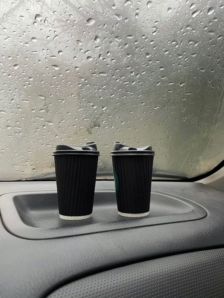 two coffee cups sitting on the dashboard of a car in front of a rain covered windshield