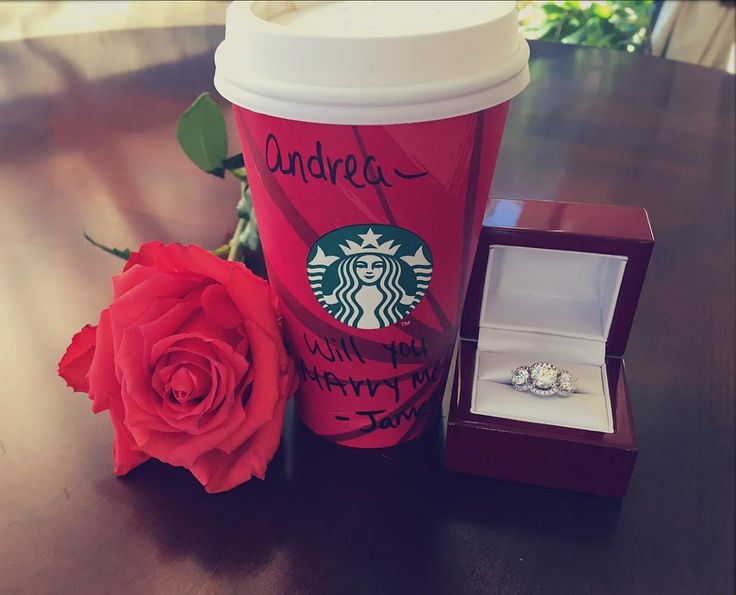 a starbucks cup with a ring on it next to a red rose and a box