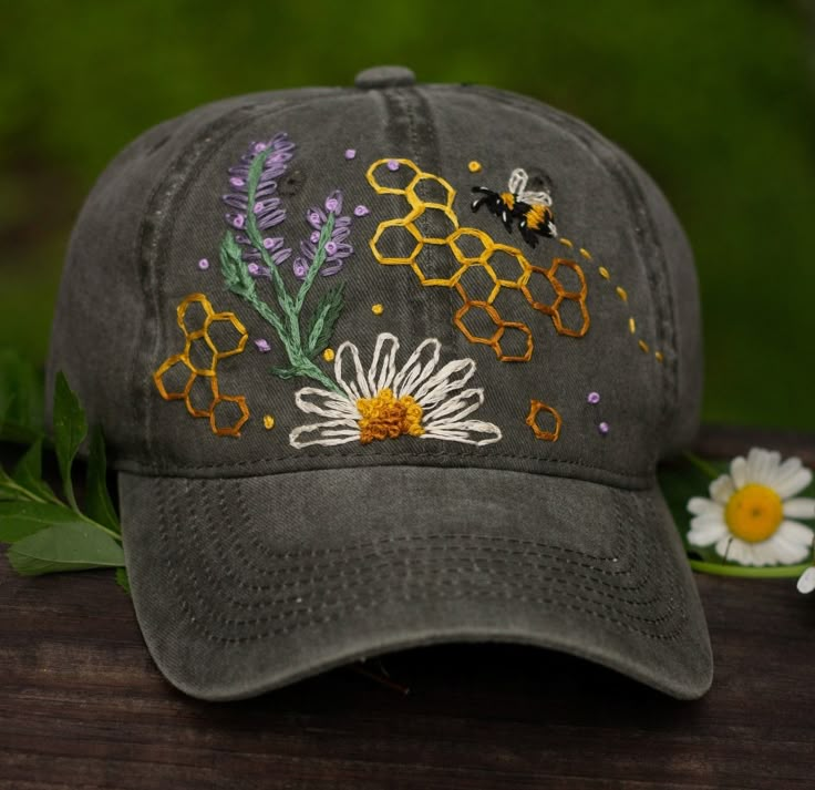 a gray hat with embroidered bees and flowers on it sitting on a wooden table next to daisies