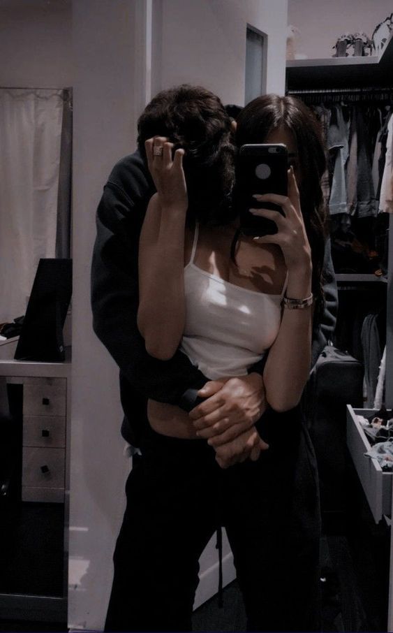 a man and woman hug in front of a mirror while looking at their cell phones