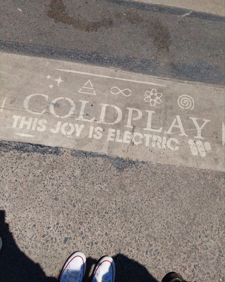 someone is standing on the sidewalk with their feet up in front of him and there is a sign that says coldplay this joy is electric