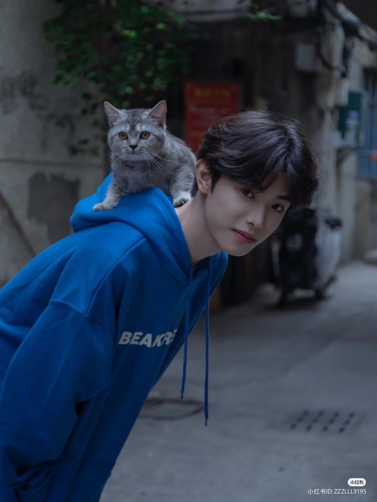 a cat sitting on the back of a man's head while he is wearing a blue hoodie