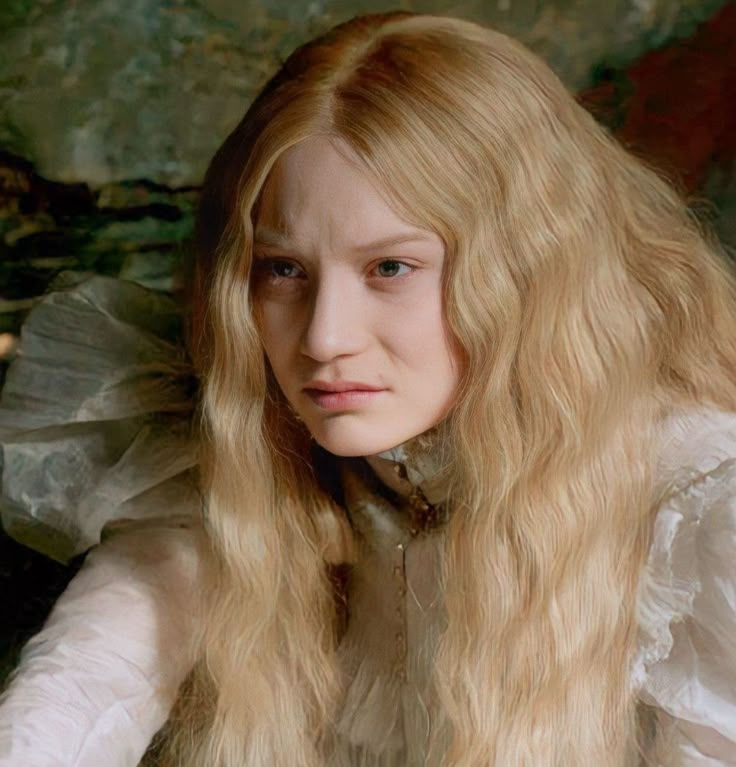 a woman with long blonde hair sitting in front of a wall and looking at the camera