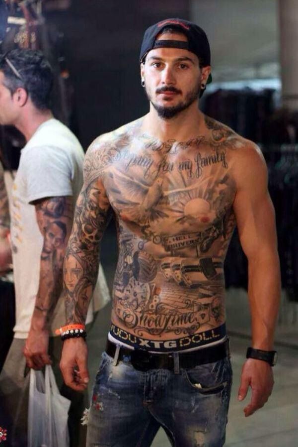 a man with lots of tattoos on his chest standing in front of other men wearing jeans and t - shirts