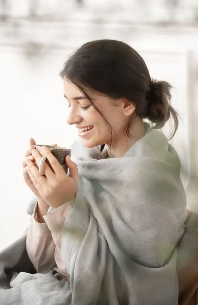 a woman sitting in a chair holding a cup and looking at her cell phone while wearing a shawl