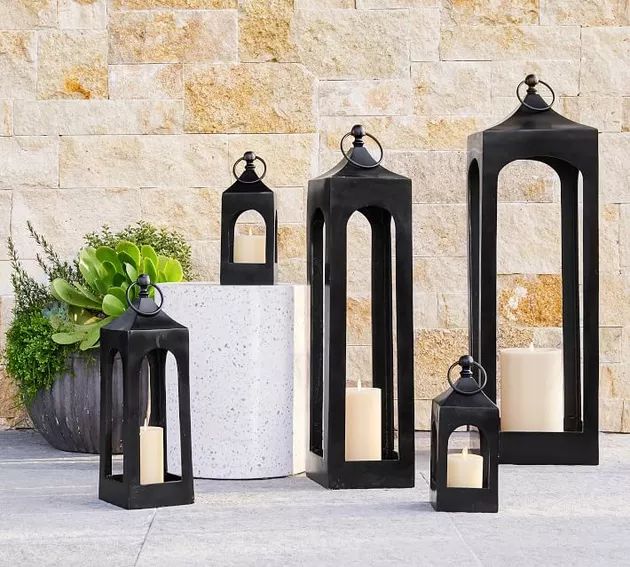 four black lanterns sitting next to each other on the ground near a stone wall and potted plant