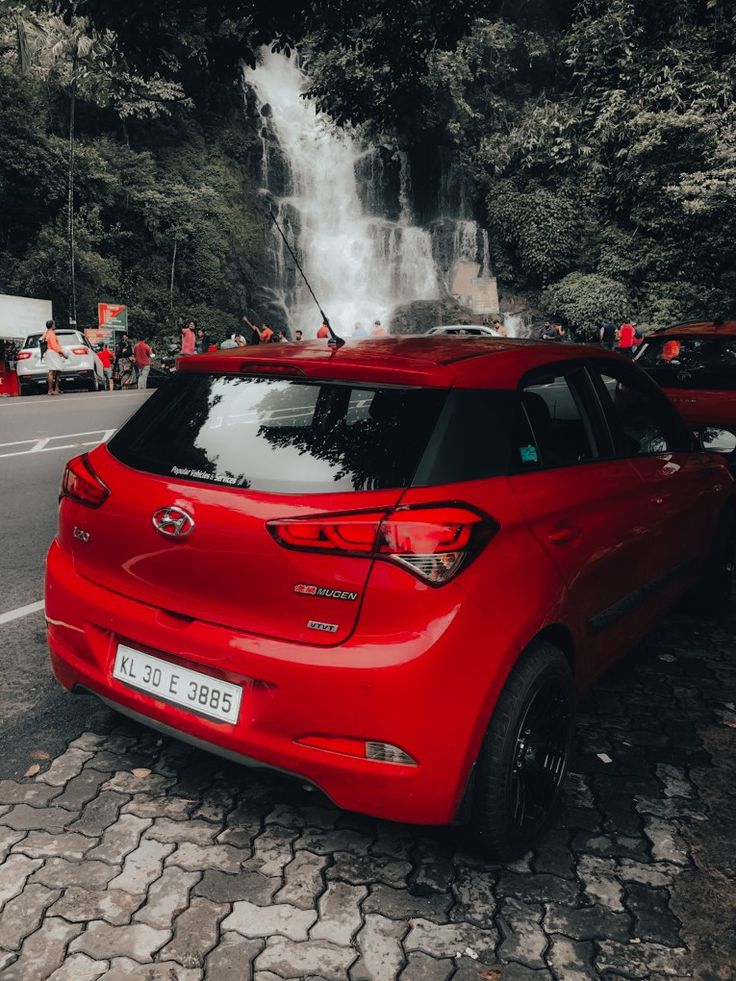 a red car parked next to a waterfall in the middle of a road with people standing around it