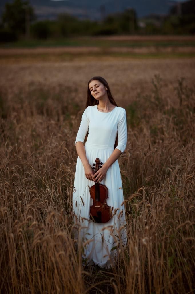 a woman in a white dress holding a violin standing in tall grass with mountains in the background