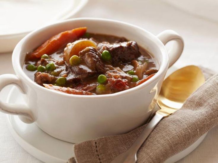 a white bowl filled with meat and vegetables on top of a plate next to a spoon