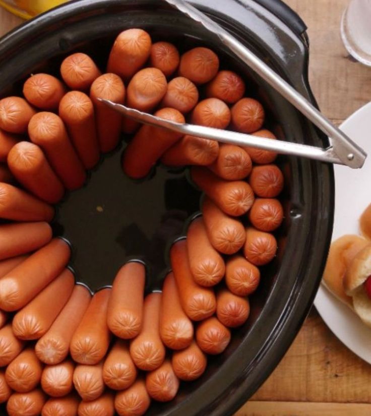 hot dogs in buns being cooked in a crock pot with tongs on the side