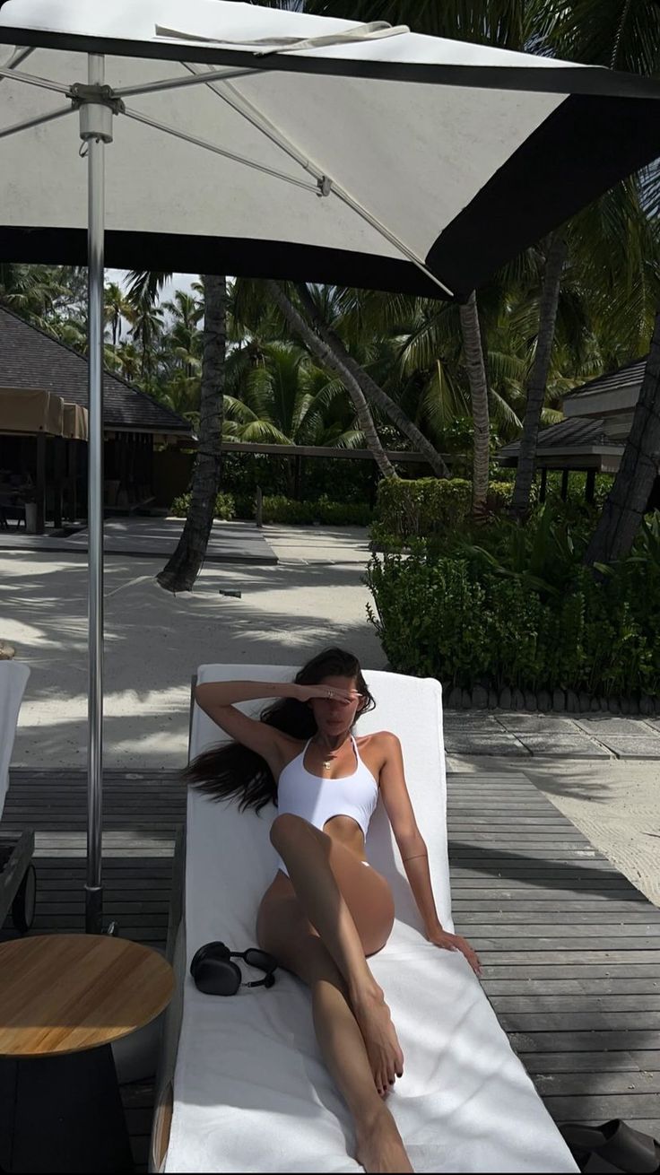 a woman in a white bathing suit sitting on a chaise lounge under an umbrella