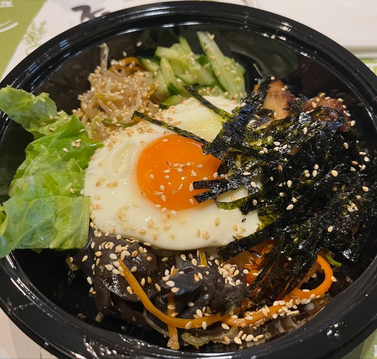 a black plastic bowl filled with vegetables and an egg on top of sesame seed sprinkles