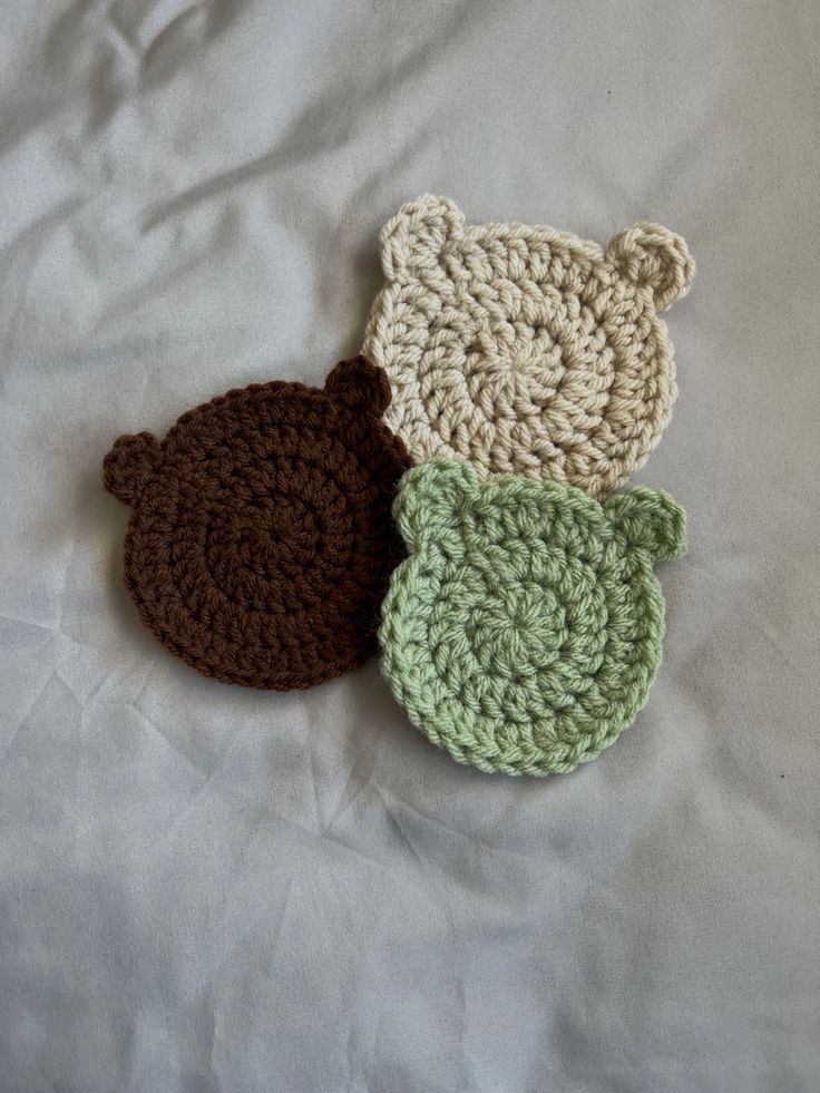 three crocheted teddy bears laying on top of a white bed sheet, one is green and the other is brown