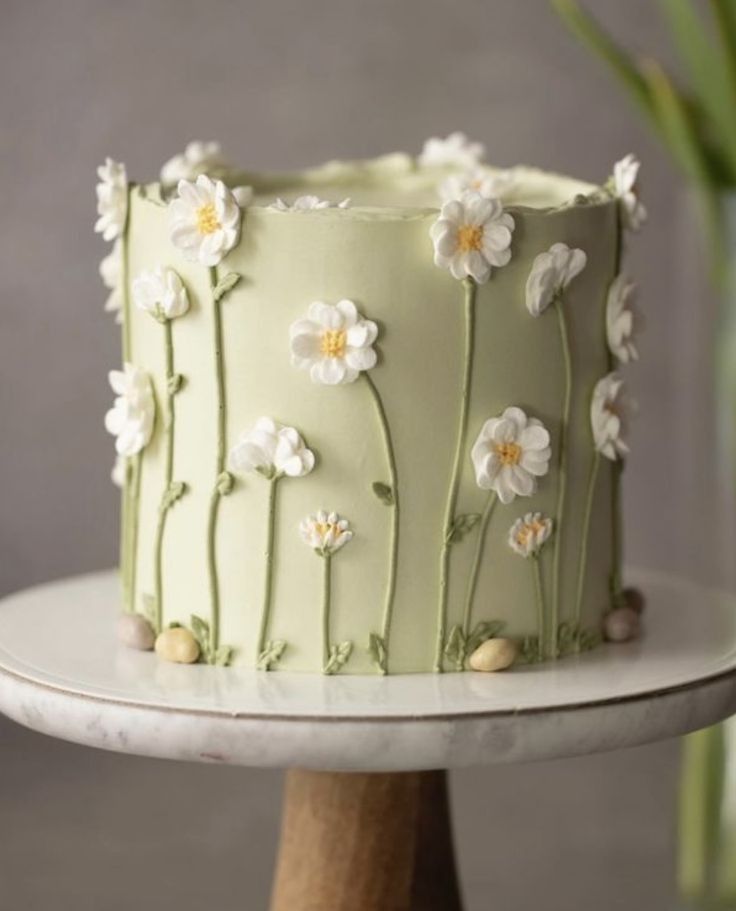 a green cake with white flowers on it sitting on top of a wooden stand next to a potted plant