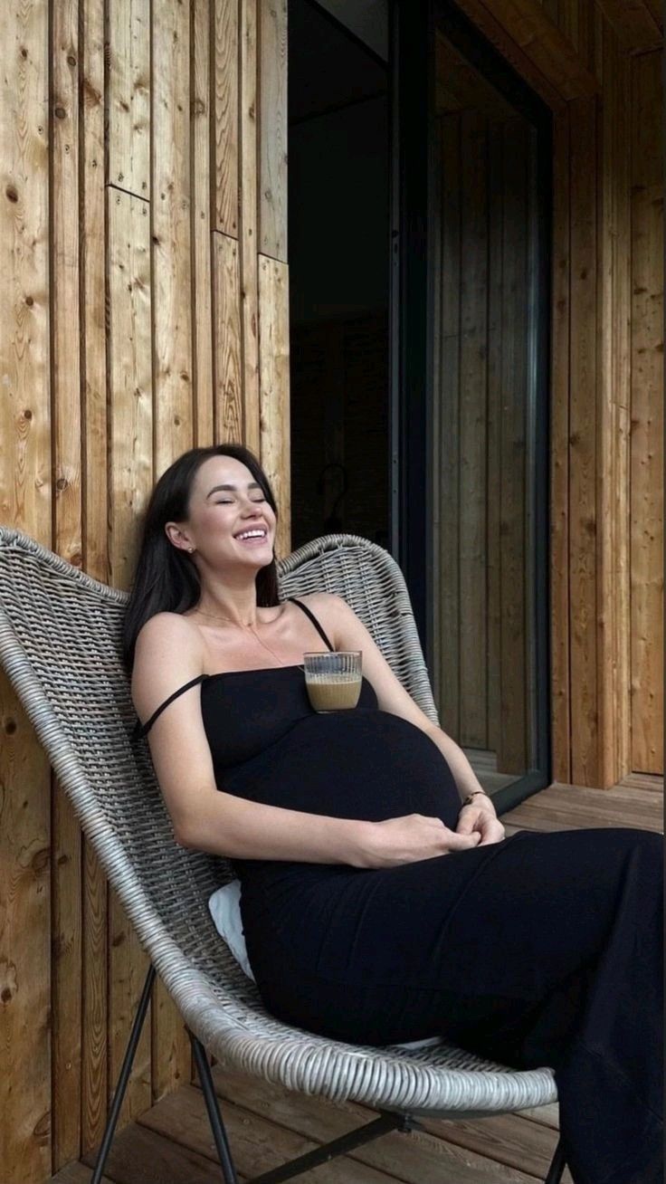 a pregnant woman sitting in a chair with a cup of coffee on her lap and smiling