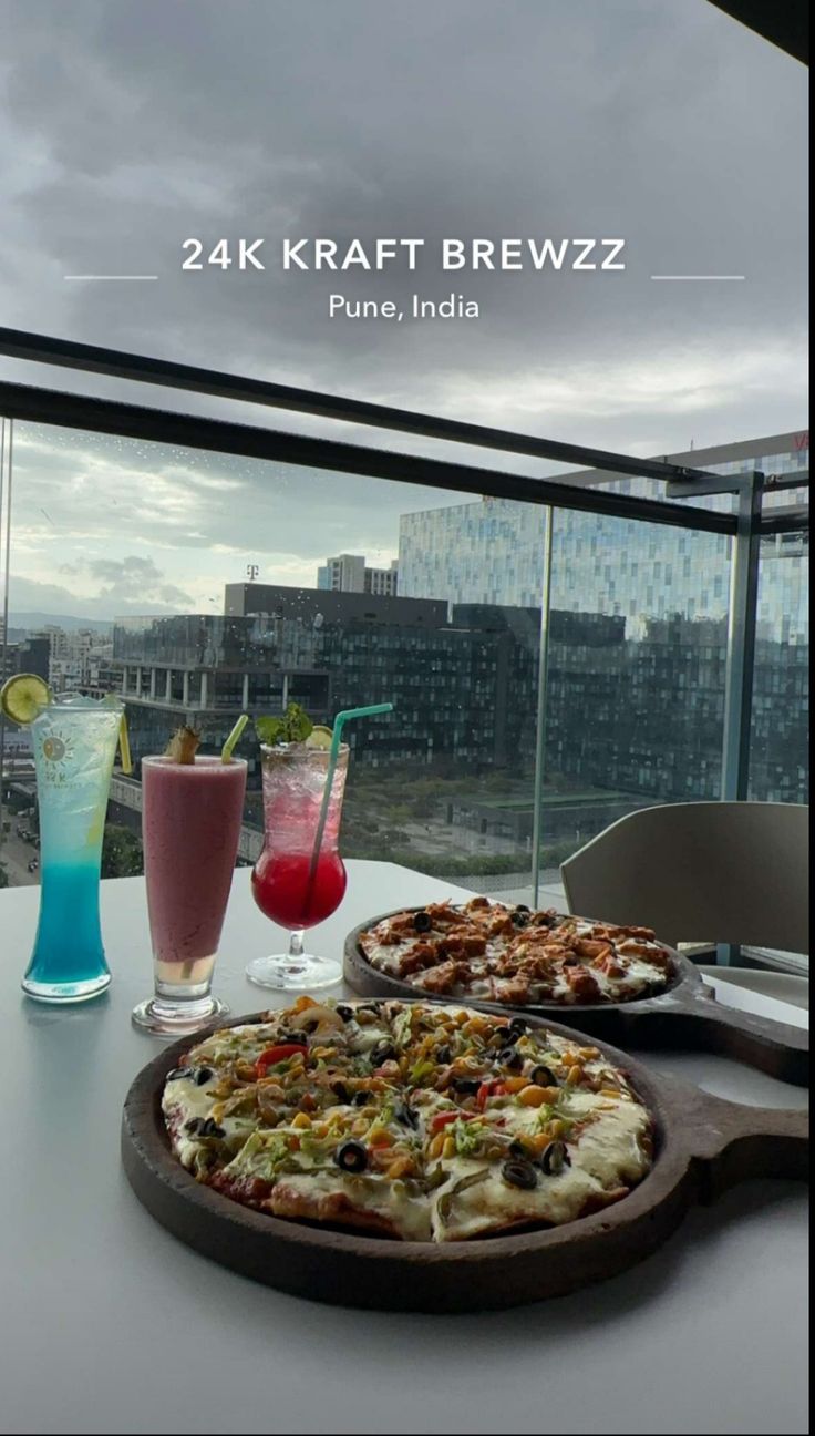 two pizzas sitting on top of a white table next to drinks and a window