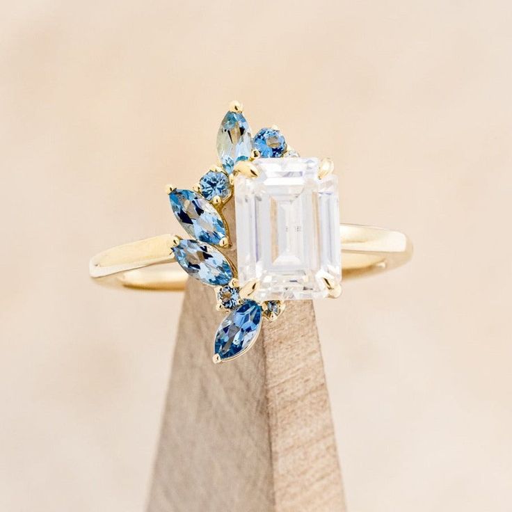 an emerald cut ring with blue and white stones on the side, sitting on top of a wooden stand