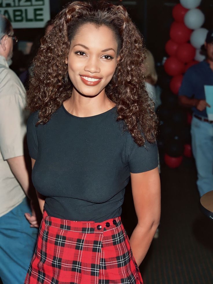 a woman with long curly hair wearing a red plaid skirt and black t - shirt