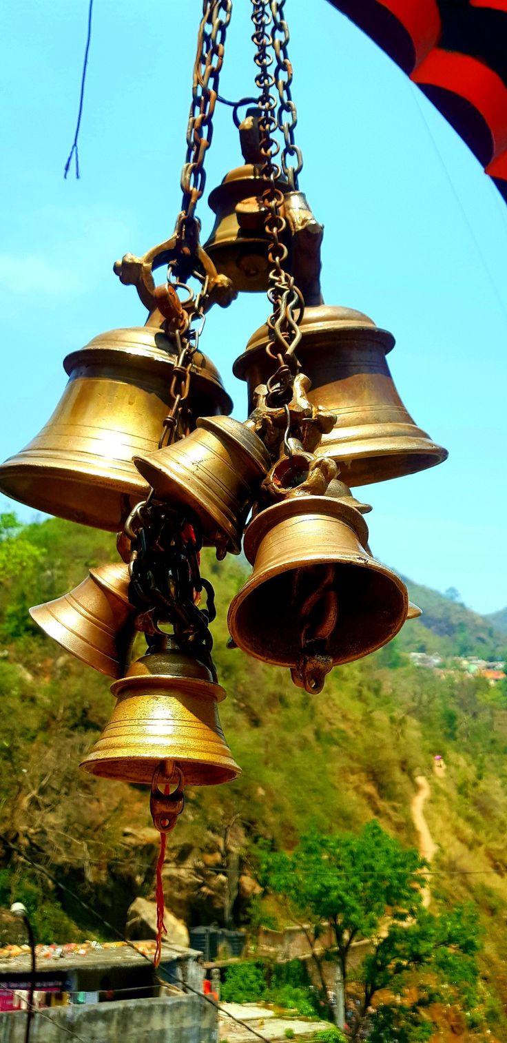 several bells hanging from a chain in front of some trees and hills with blue sky