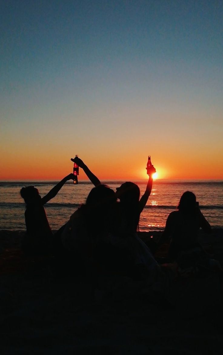 two people are sitting on the beach watching the sun go down and one person is holding up a bottle