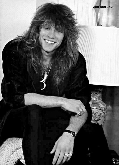 a black and white photo of a man with long hair sitting on a bed smiling