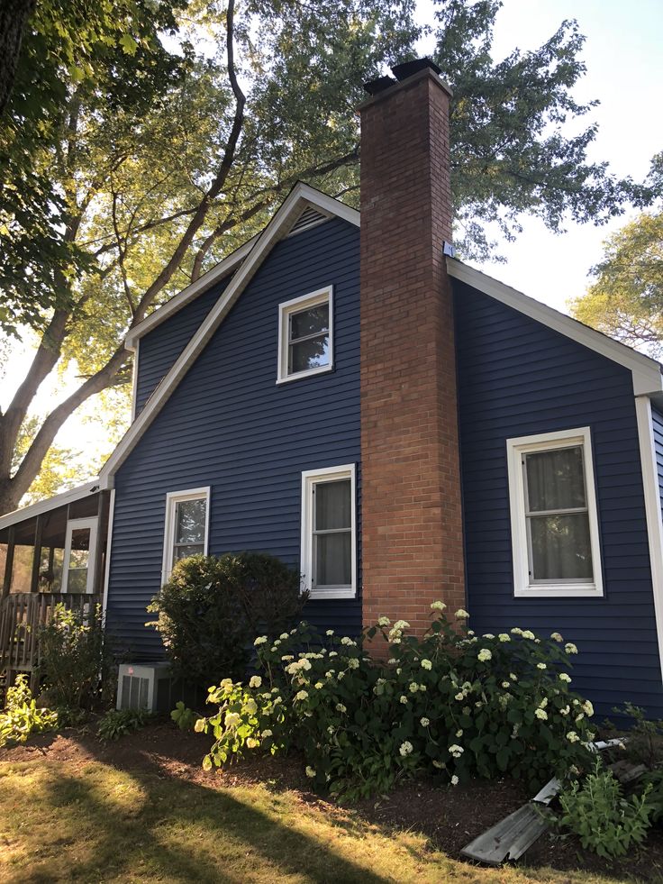 a blue house with white windows and a brick chimney in the front yard is shown