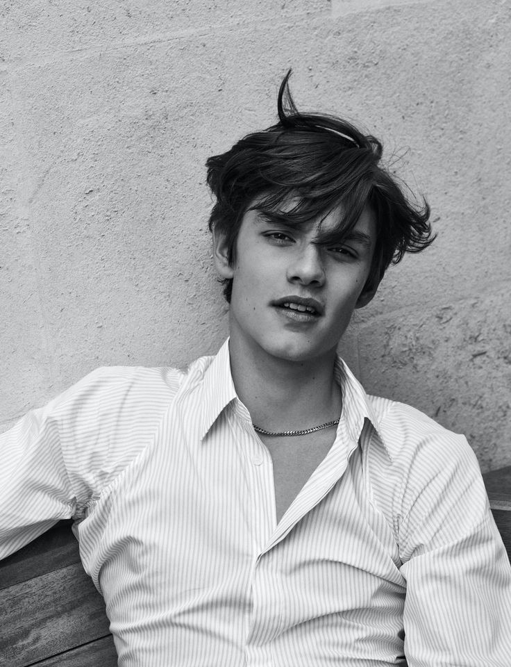 a young man sitting on top of a wooden bench next to a wall and wearing a shirt