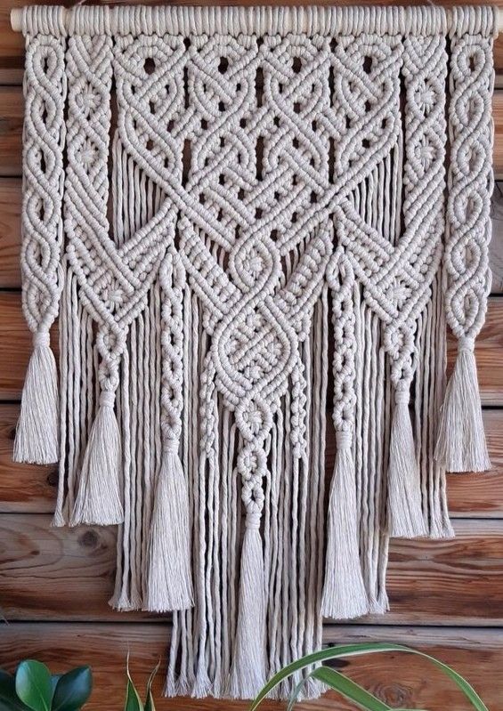 macrame wall hanging with tassels and beads on a wooden wall next to a potted plant