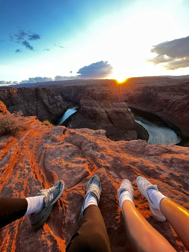 two people are sitting on the edge of a cliff looking out over a river at sunset