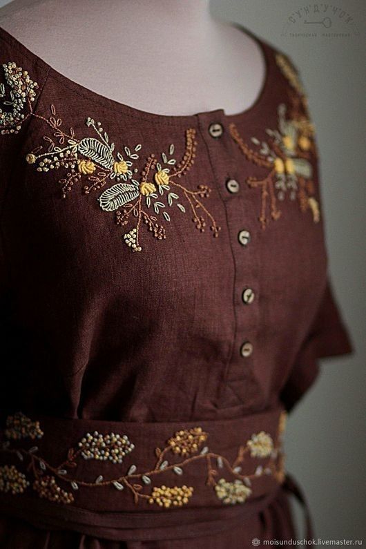 a woman wearing a brown dress with flowers on it's chest and sleeves, standing in front of a white wall