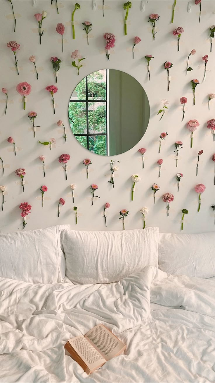 a bed with white sheets and pink flowers on the wall next to a round mirror