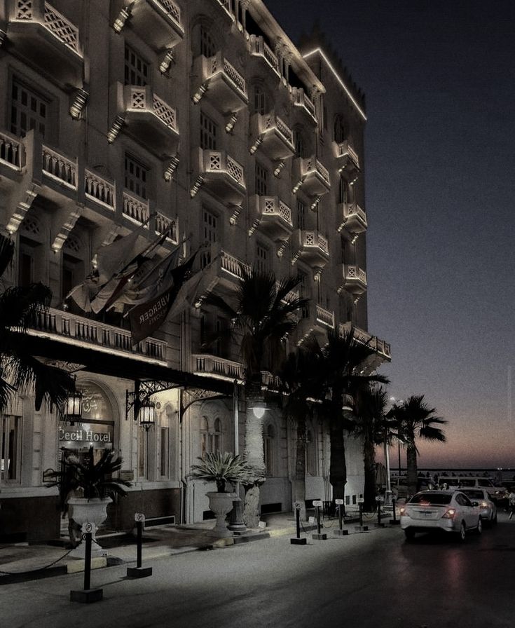 an old building is lit up at night with palm trees in the foreground and cars parked on the street