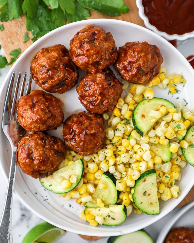 a white plate topped with meatballs next to corn and cucumber on a table