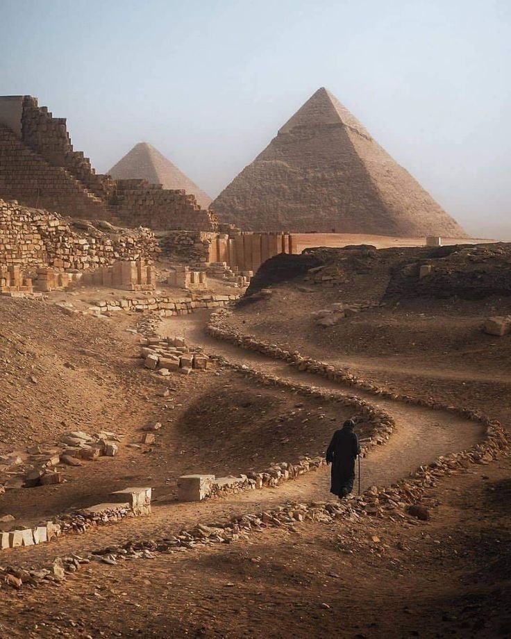 a man walking down a dirt path in front of three pyramids