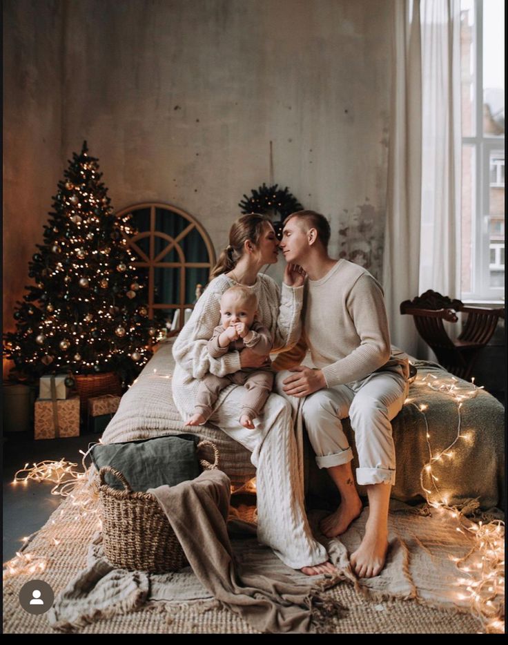 a man and woman are sitting on a bed with their baby in front of a christmas tree