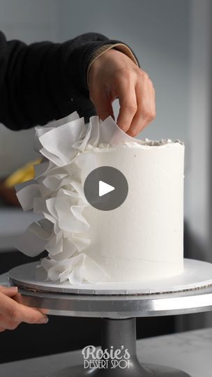 a woman is decorating a white cake with ruffles on it while another person holds out their hand