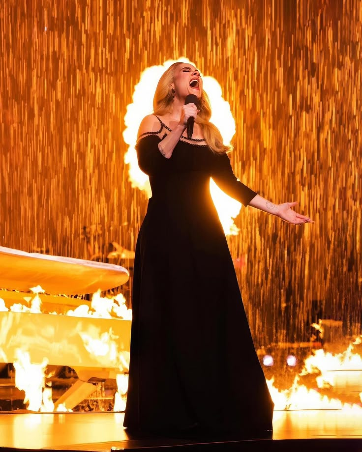 a woman in a black dress singing on stage with fire and water behind her,