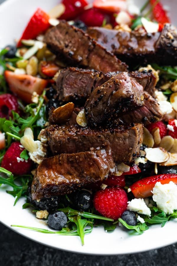 steak salad with strawberries, blueberries, and nuts on a white plate sitting on a black table